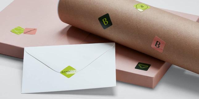 Enveloppe blanche fermée par un autocollant carré vert, boîte rose et tube postal en carton décoré de mini autocollants