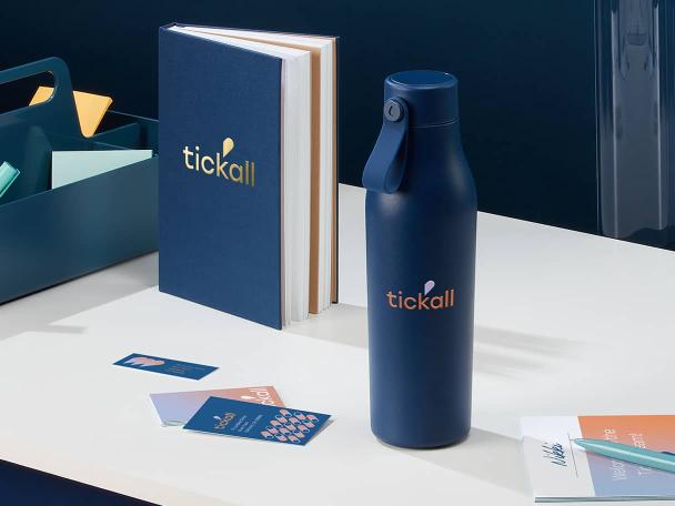 Custom blue water bottle with logo, blue hardcover notebook, branded marketing materials, pen and a blue desk storage basket on a table