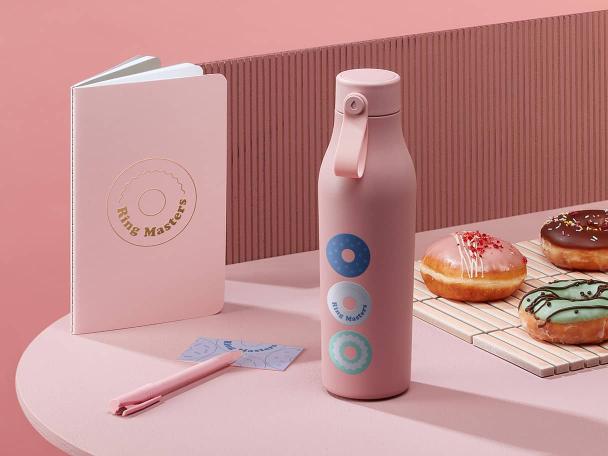 Pink custom bottle with a doughnut company logo next to a branded pink softcover notebook, a pen, a blue business card and 3 doughnuts 