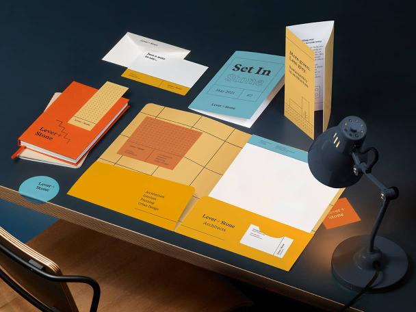 Escritorio negro con lámpara y colección de materiales impresos en naranja, azul y amarillo, que incluyen tarjetas de felicitación, cuadernos personalizados, pegatinas y volante doblado.