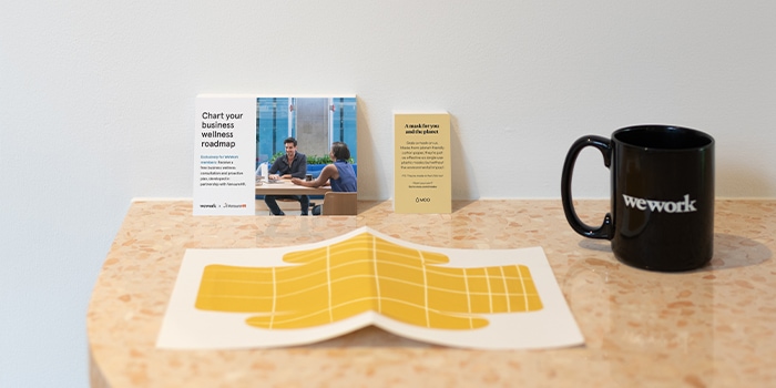Yellow paper face mask design on a desk with MOO x WeWork postcard and minicard, and black WeWork mug