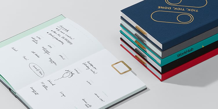 Pile of custom planners in various colors and undated planner open to the weekly spread with handwritten notes