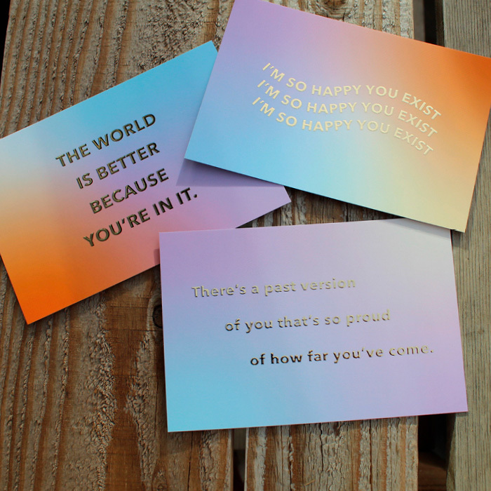 Three different postcards with encouraging phrases on them in shiny gold foil