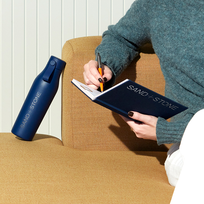 Person writing on customized Notebook. Matching Customized Water Bottle resting on arm chair.