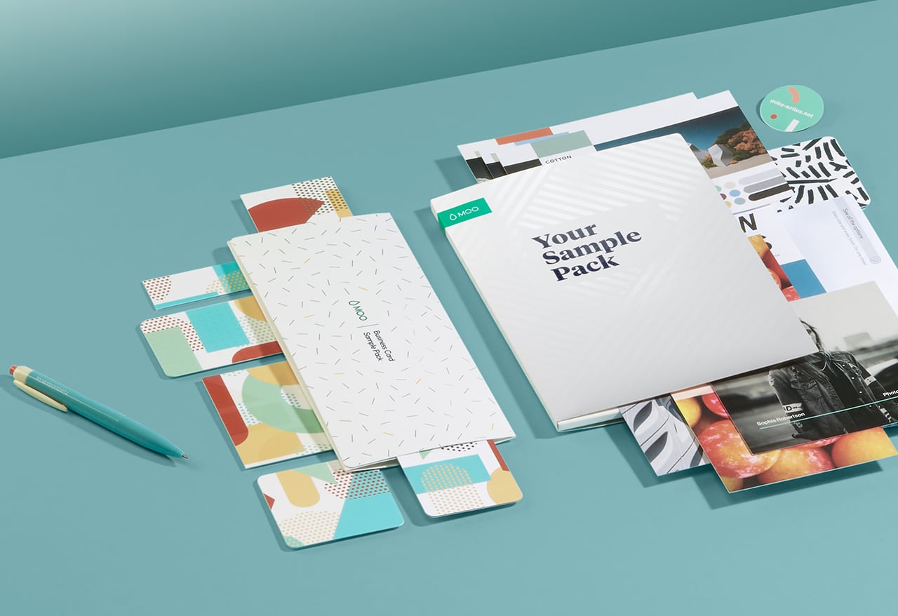 MOO-Visitenkarte und Alle Produkte Sample Packs auf blauem Tisch neben grauen Stuhl