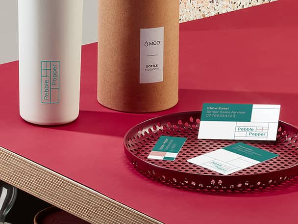 Botella de agua personalizada con logo junto a su empaque de tubo y 3 tamaños de tarjetas de visita en un plato clave rojo sobre una mesa roja