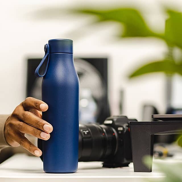 Hand, die eine dunkelblaue Wasserflasche hält Es gibt eine Kamera und einen Druck im Hintergrund.