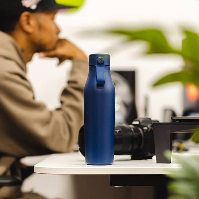 Bottiglia di bevanda blu in primo piano. Uomo seduto alla sua scrivania e fotocamera professionale in background.