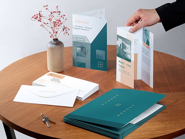 White desk and collection of print materials in orange, blue and yellow, including greeting cards, custom notebooks, stickers, folded flyer, presentation folder and water bottle.