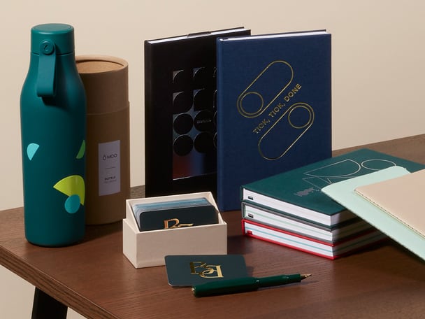 Une sélection de produits MOO sur un bureau, dont une bouteille d\'eau personnalisée, deux carnets personnalisés, une boîte de présentation contenant des cartes de visite et deux journaux à couverture souple.