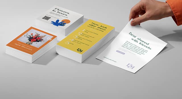 3 piles de flyers pour un service de livraison de fleurs de différentes tailles et une main prenant un grand dépliant blanc