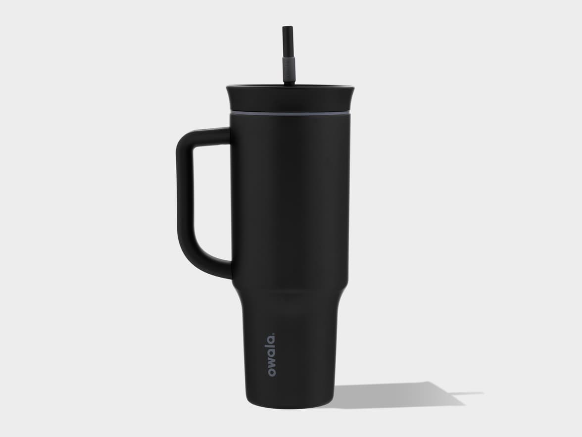 A black Owala tumbler with a handle and a straw stands upright, casting a shadow to its right, against a plain white background.