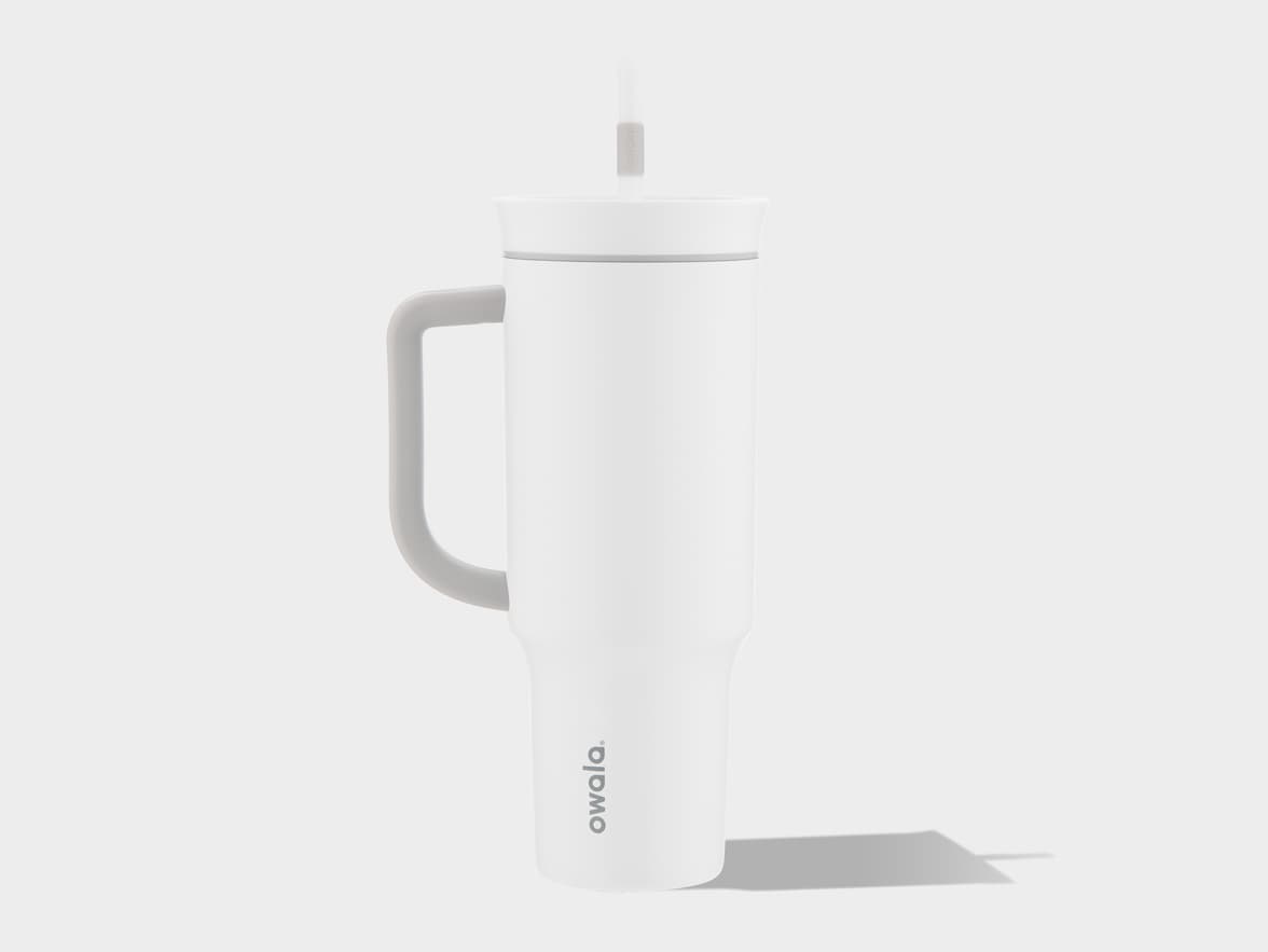 A white Owala tumbler with a gray handle and straw stands on a white surface, casting a subtle shadow to its right.