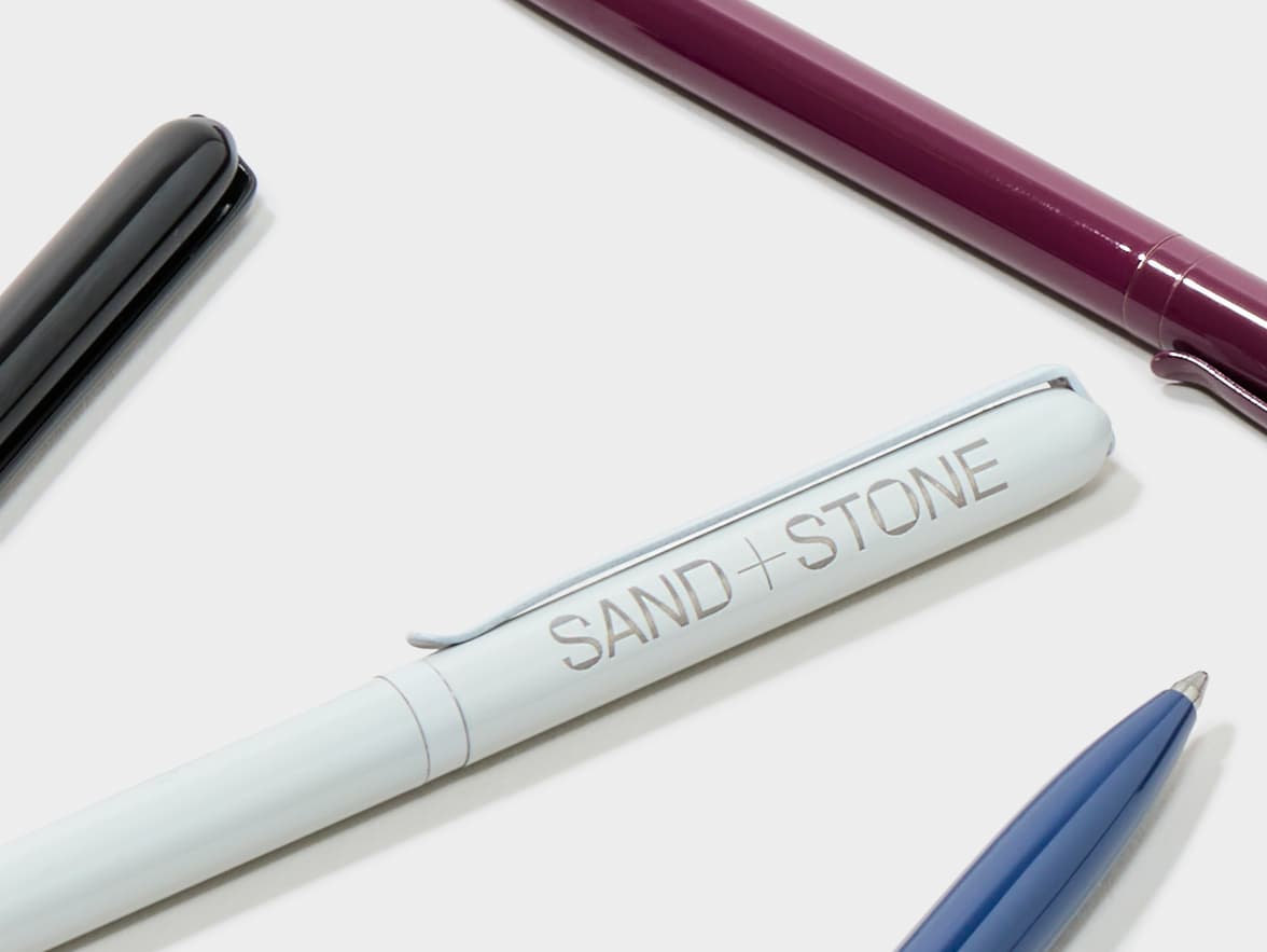Four pens, including a white pen with "SAND + STONE" branding, a black pen, a dark blue pen, and a maroon pen, lie on a white surface.