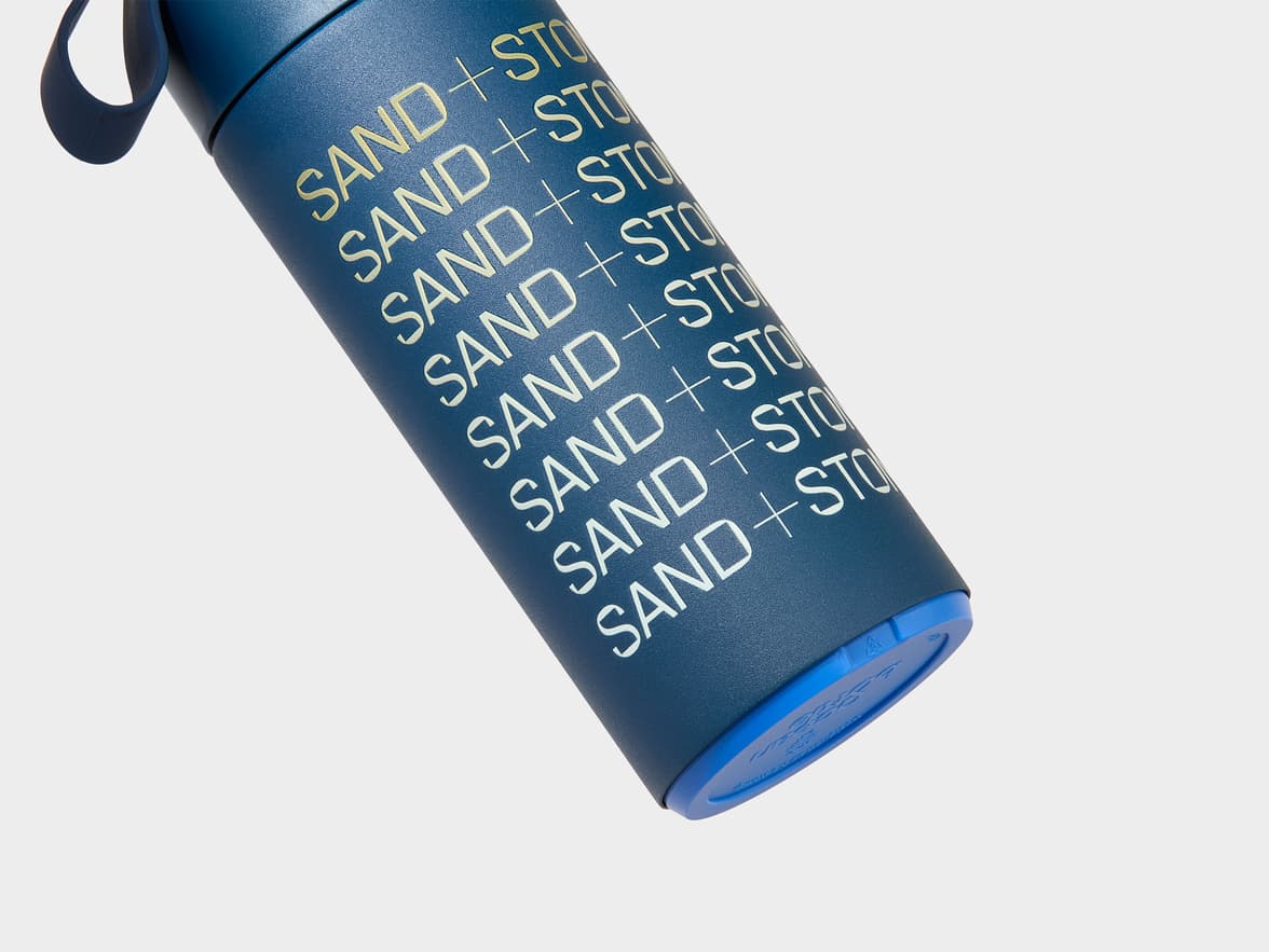 Dark blue water bottle with repeating 'SAND + STO' branding in light gold.