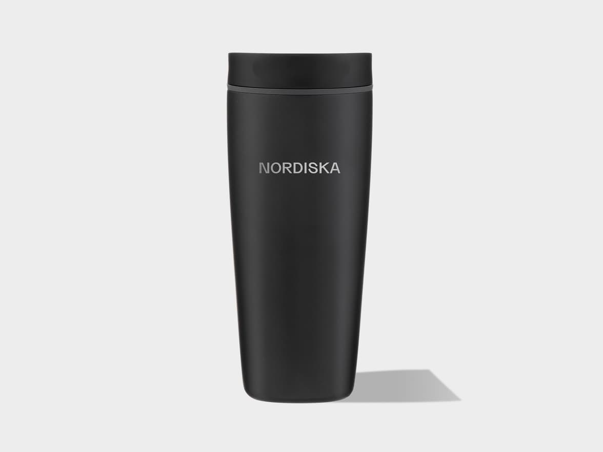 A black matte travel mug with a matching cap and a thin gray band below it. The word "NORDISKA" is printed vertically in light gray on the front. The mug casts a shadow to its right on a light gray background.