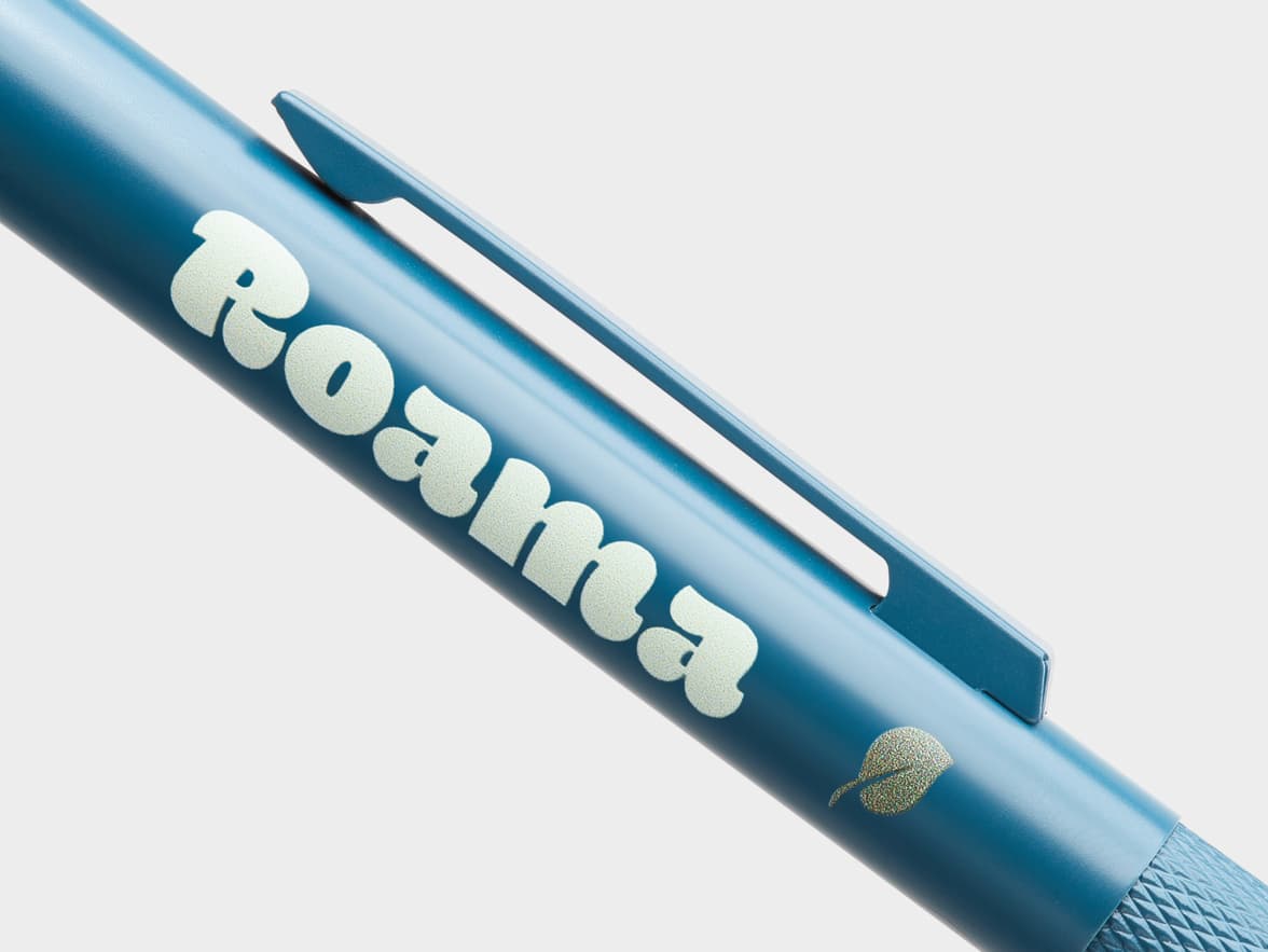 A close-up of a blue pen with "Roama" branded on its side, next to a small leaf logo.