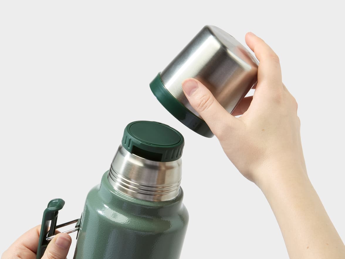 A hand removing the stainless steel cup from a green thermos held by another hand.