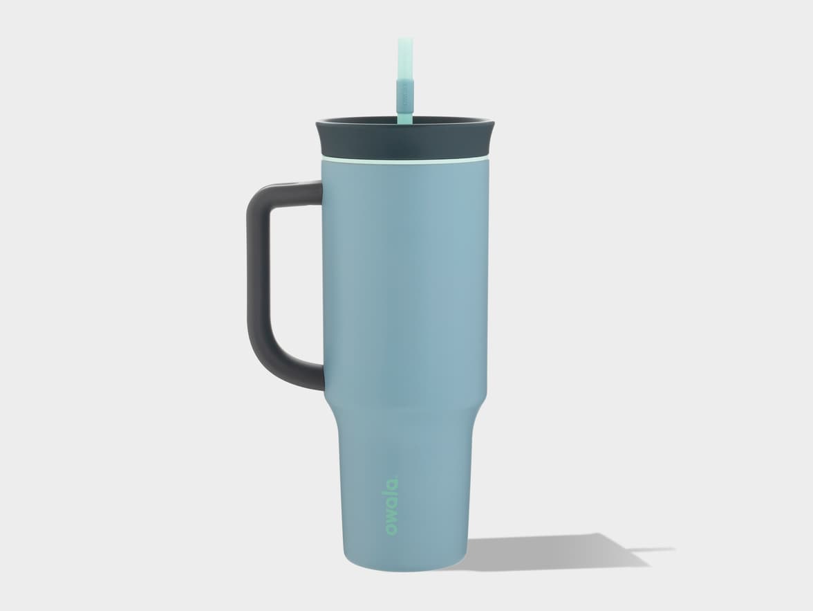 A light blue Owala tumbler with a dark gray handle and lid, featuring a light green straw and light green "owala." branding on the side, standing on a light gray surface with a shadow.