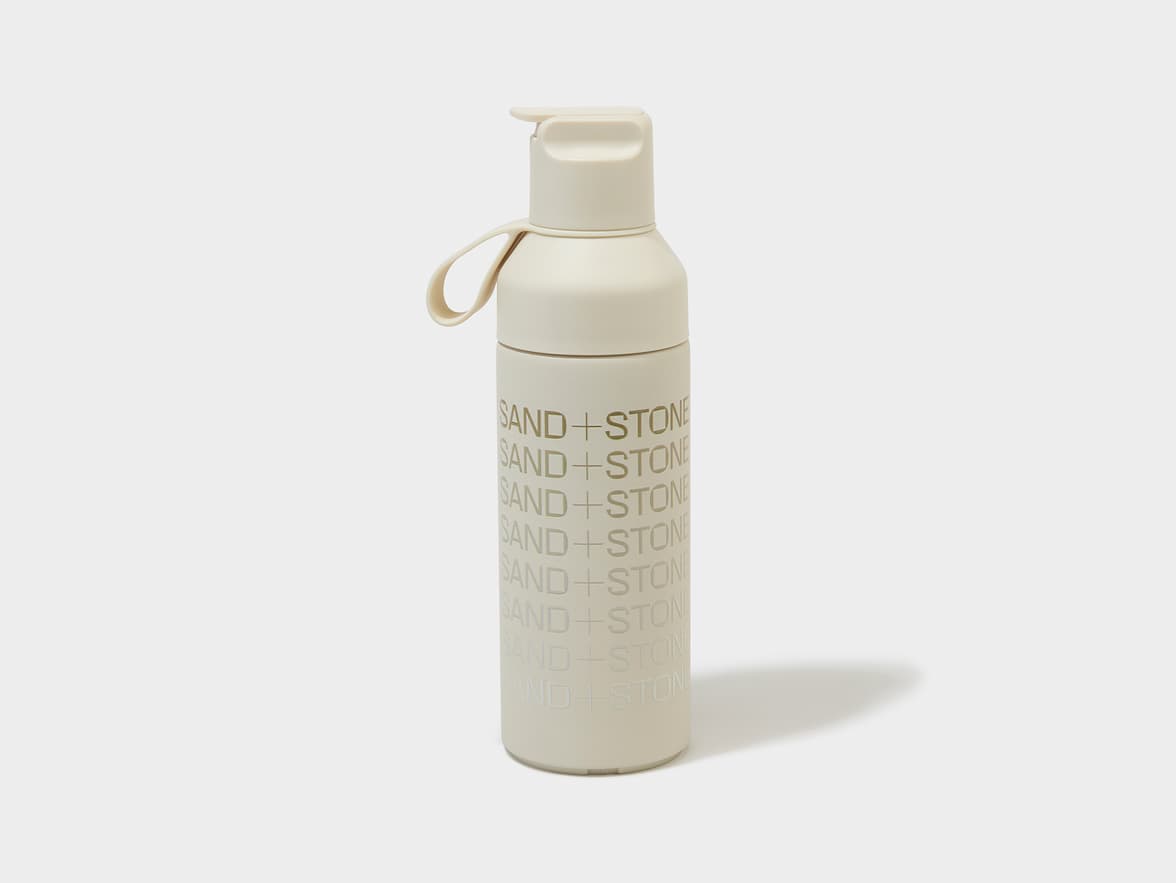 A cream-colored reusable water bottle with "SAND+STONE" branding repeated vertically on its side.