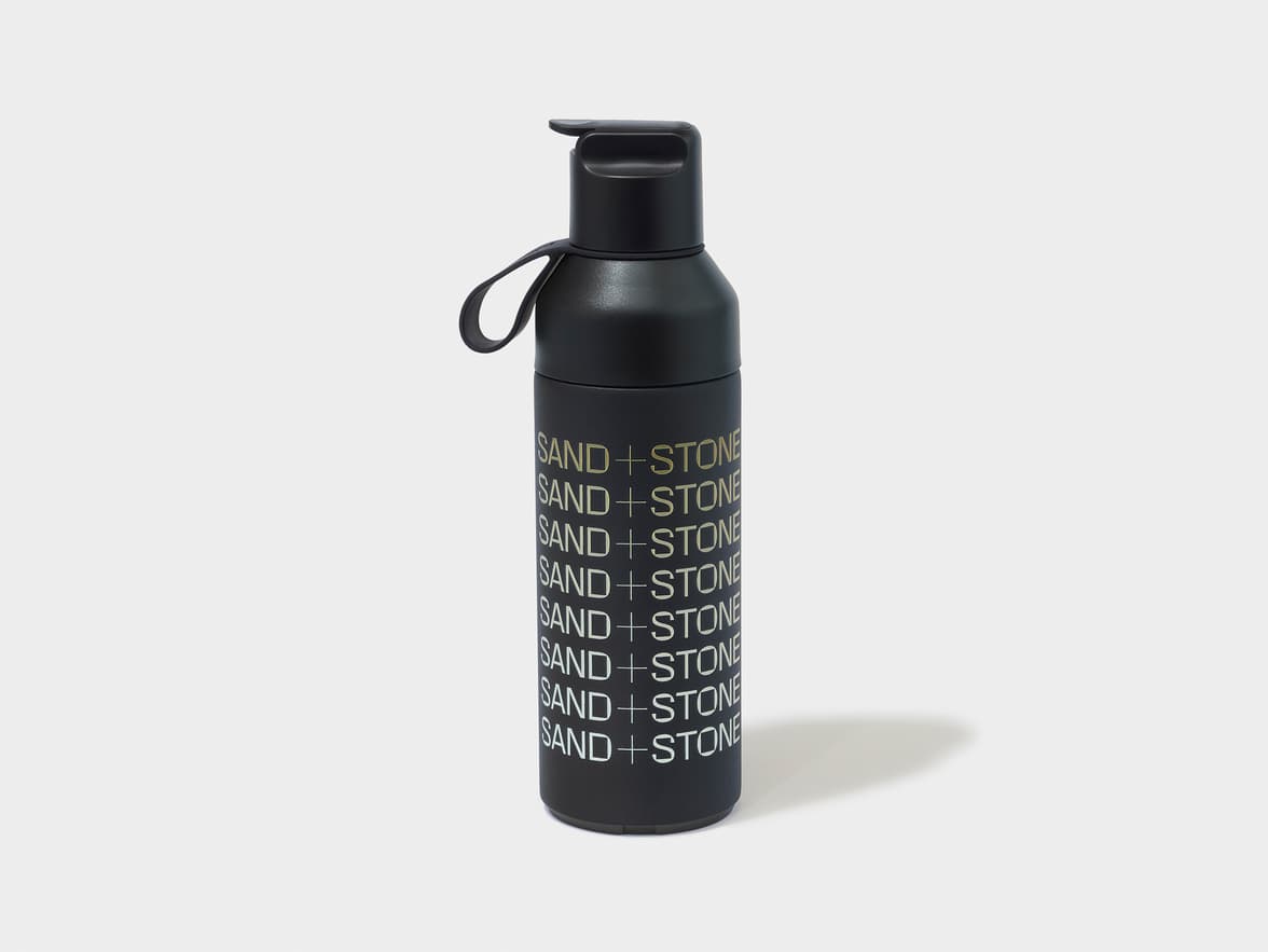Black water bottle with the branding "SAND + STONE" repeated vertically in gold lettering.