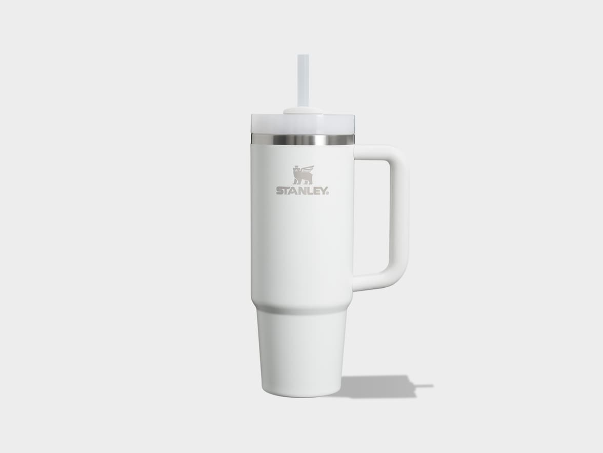 A white Stanley Quencher FlowState H2.0 tumbler, shown upright and centered, with a clear straw extended from its top. The mug has a silver band below the lid and a large white handle on its right side. The Stanley logo is embossed on the front.