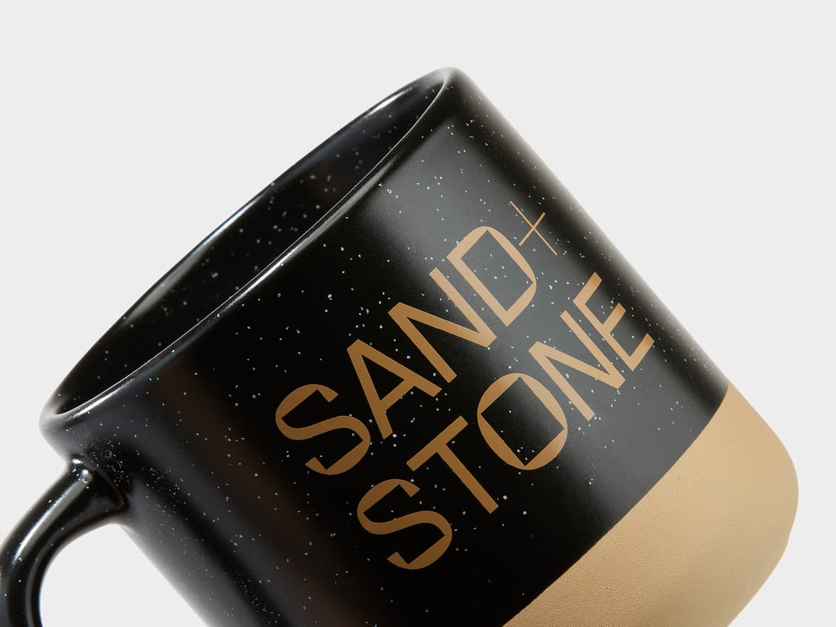 Black speckled ceramic mug with a tan base, branded "SAND + STONE".
