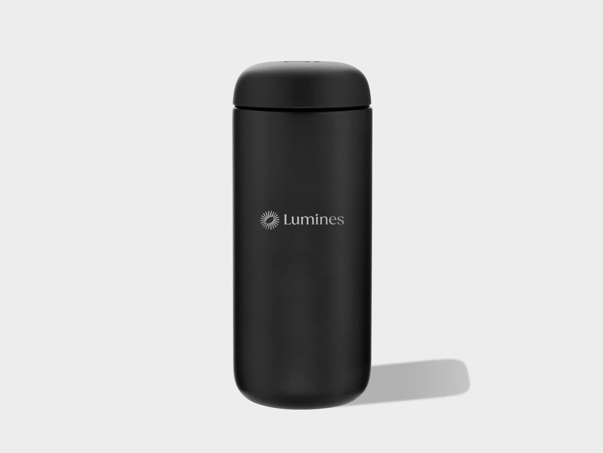 A black matte cylindrical travel mug or thermos with a rounded top and bottom stands vertically on a light grey background. A silver "Lumines" logo, accompanied by a stylized sunburst icon, is centered on the upper front of the mug.