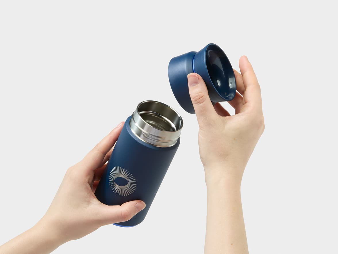 A pair of hands opens a dark blue thermos with a silver logo featuring an oval center and radiating lines, revealing its stainless steel interior.