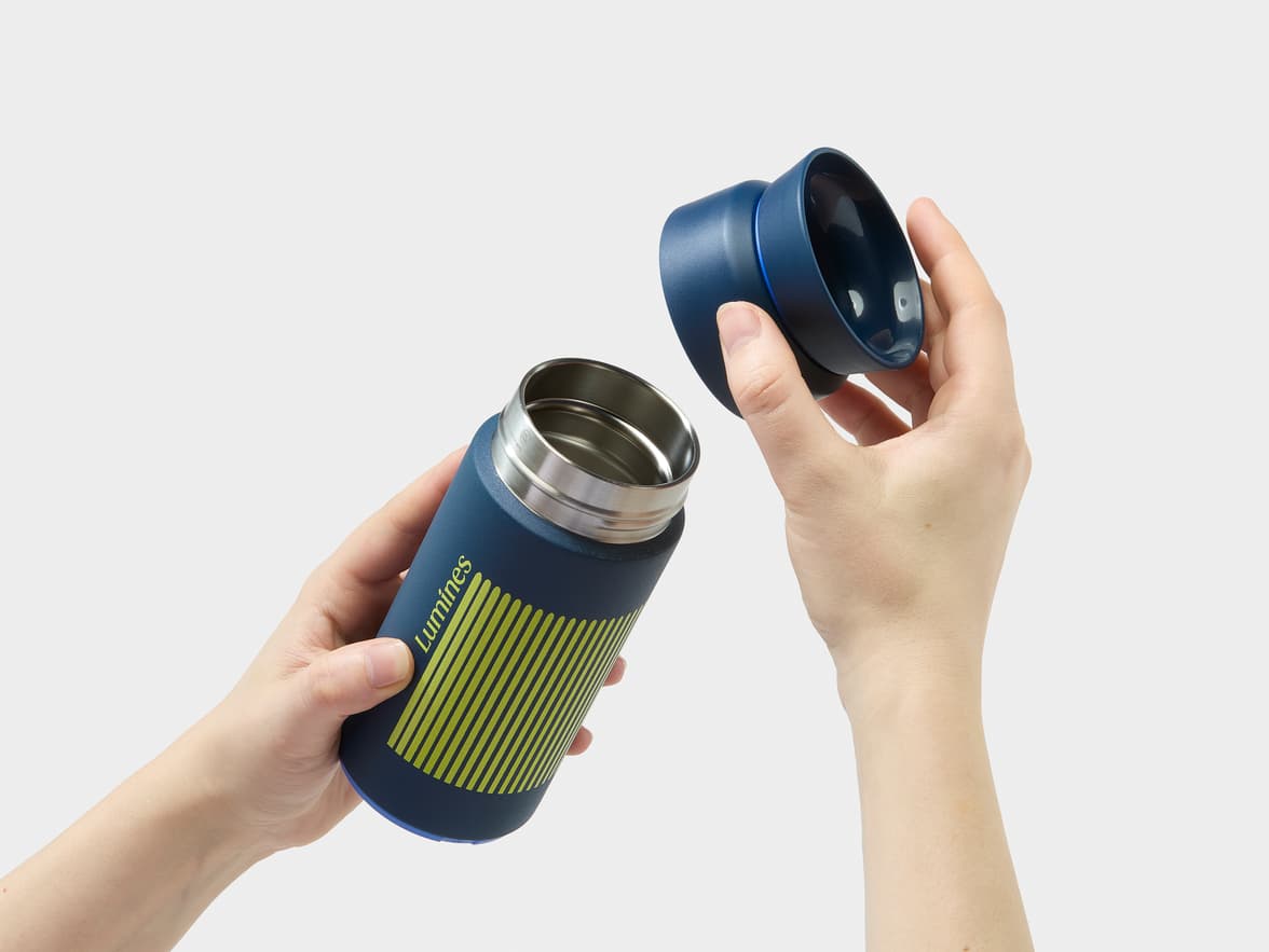 A person's hands hold a dark blue "Luminēs" brand thermos with green stripes, open with the lid removed.