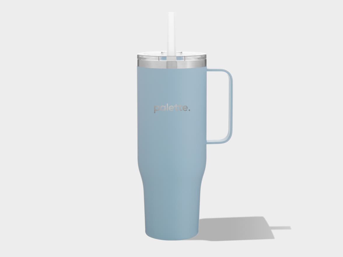 A light blue matte insulated tumbler with a handle and a clear lid with a straw, featuring a silver "palette." logo, stands upright on a light gray background with a shadow cast to the right.
