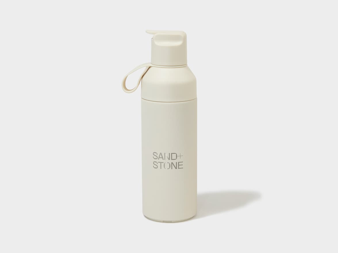 A cream-colored water bottle with a loop handle and a flip-top lid, branded "SAND+ STONE".