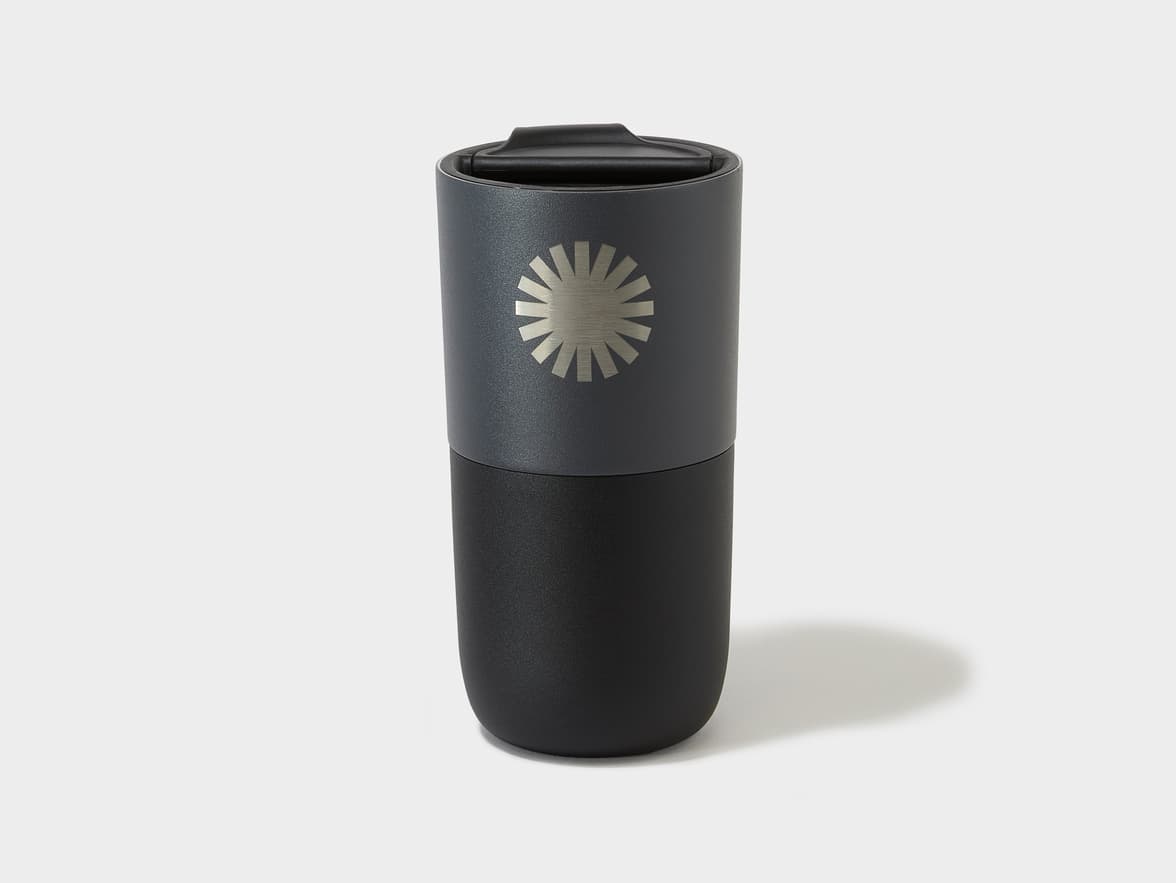 A two-toned dark grey and black insulated tumbler with a silver sunburst logo on the dark grey section.