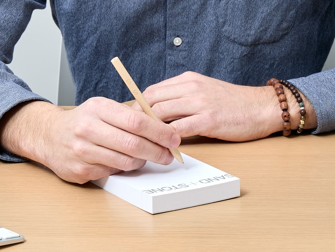 A person in a denim shirt writes on a white notepad labeled "SAND + STONE" with a light brown pencil.