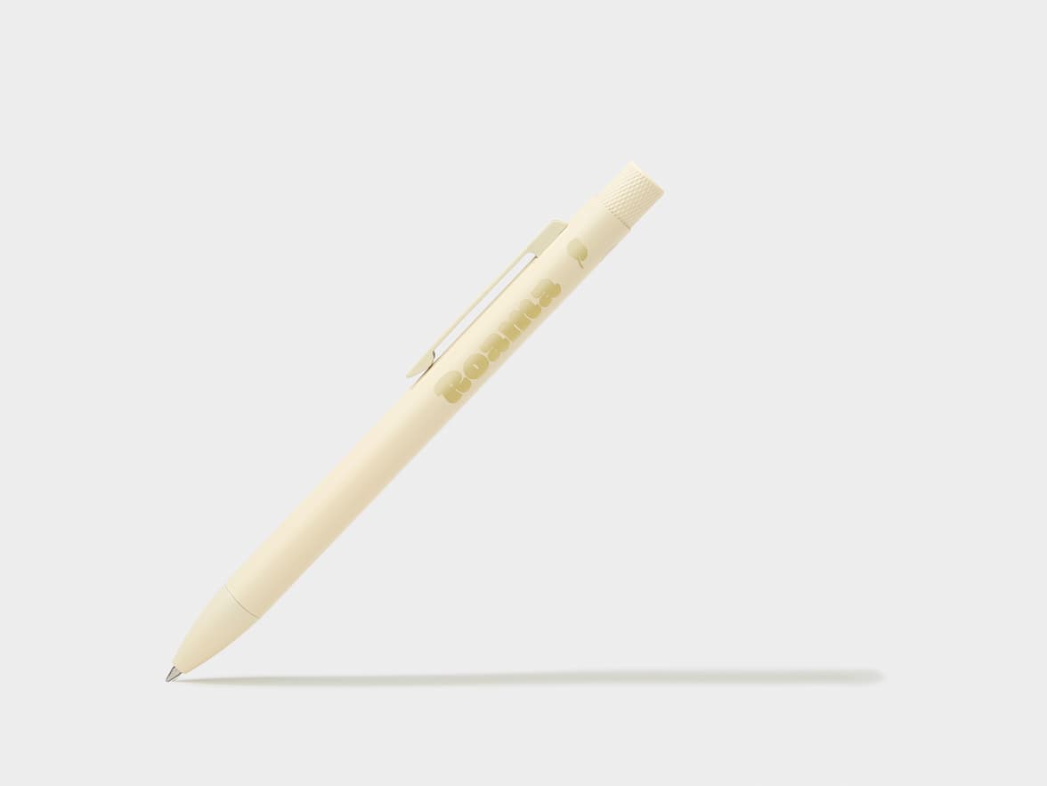 A light beige ballpoint pen with "Roama" branding on its barrel and a textured grip, with its tip extended, casting a shadow on a white background.