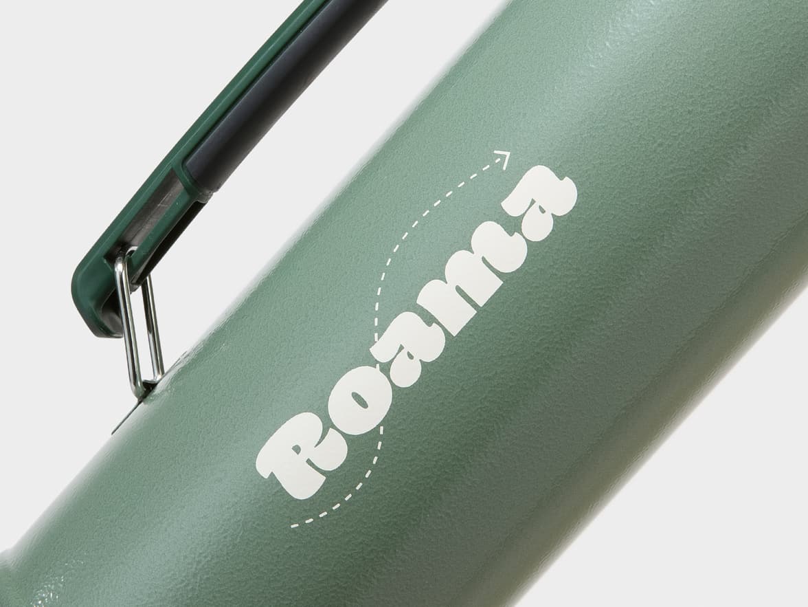 A close-up of a green insulated bottle with a handle, featuring the white 'Roama' brand logo and a dashed arrow.