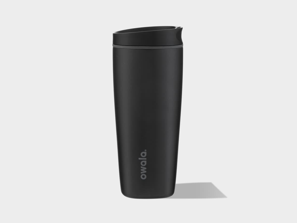 A black Owala brand travel mug with a black lid and a dark grey accent ring stands upright on a light grey background, casting a shadow to the right. The "owala" logo is subtly visible near the bottom of the mug.