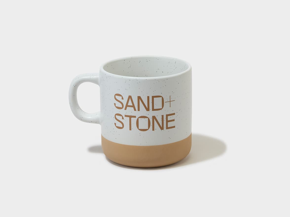 A white speckled mug with a natural brown base and a handle, featuring the branding "SAND + STONE" in brown text.
