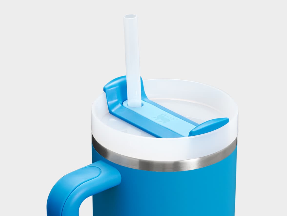 A close-up shot of a blue insulated mug with a white lid, a blue handle, and a clear plastic straw inserted into a blue, winged mechanism on the lid. A subtle logo is embossed on the blue part of the lid.