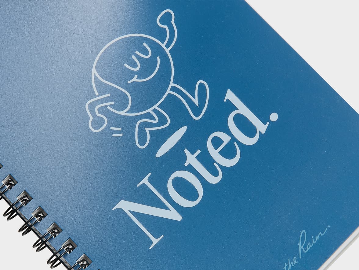 Blue spiral notebook with a white illustration of a happy character, the text "Noted.", and "the Rain®" branding.