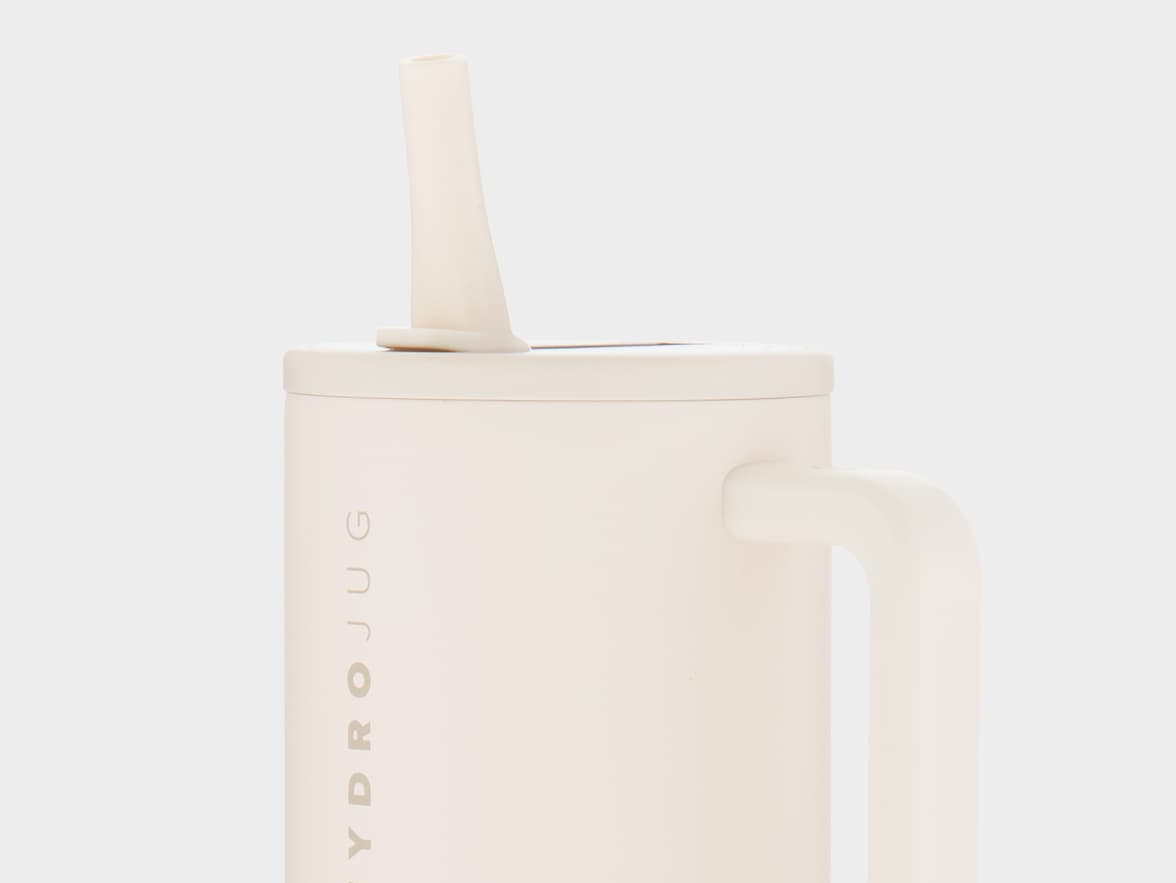 A close-up, cropped view of a cream or light beige "HydroJug" water bottle against a plain light grey background. The jug has a matching lid with a straw extending upwards from its center. A handle of the same color is visible on the right side. The brand name "HYDROJUG" is written vertically in a slightly darker, muted tone on the left side of the jug.