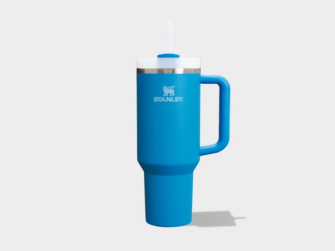 A bright blue Stanley Quencher H2.0 FlowState Tumbler with a handle and a clear straw protruding from its white lid, casting a shadow on a light grey background. The silver Stanley bear logo and wordmark are visible on the front of the mug.