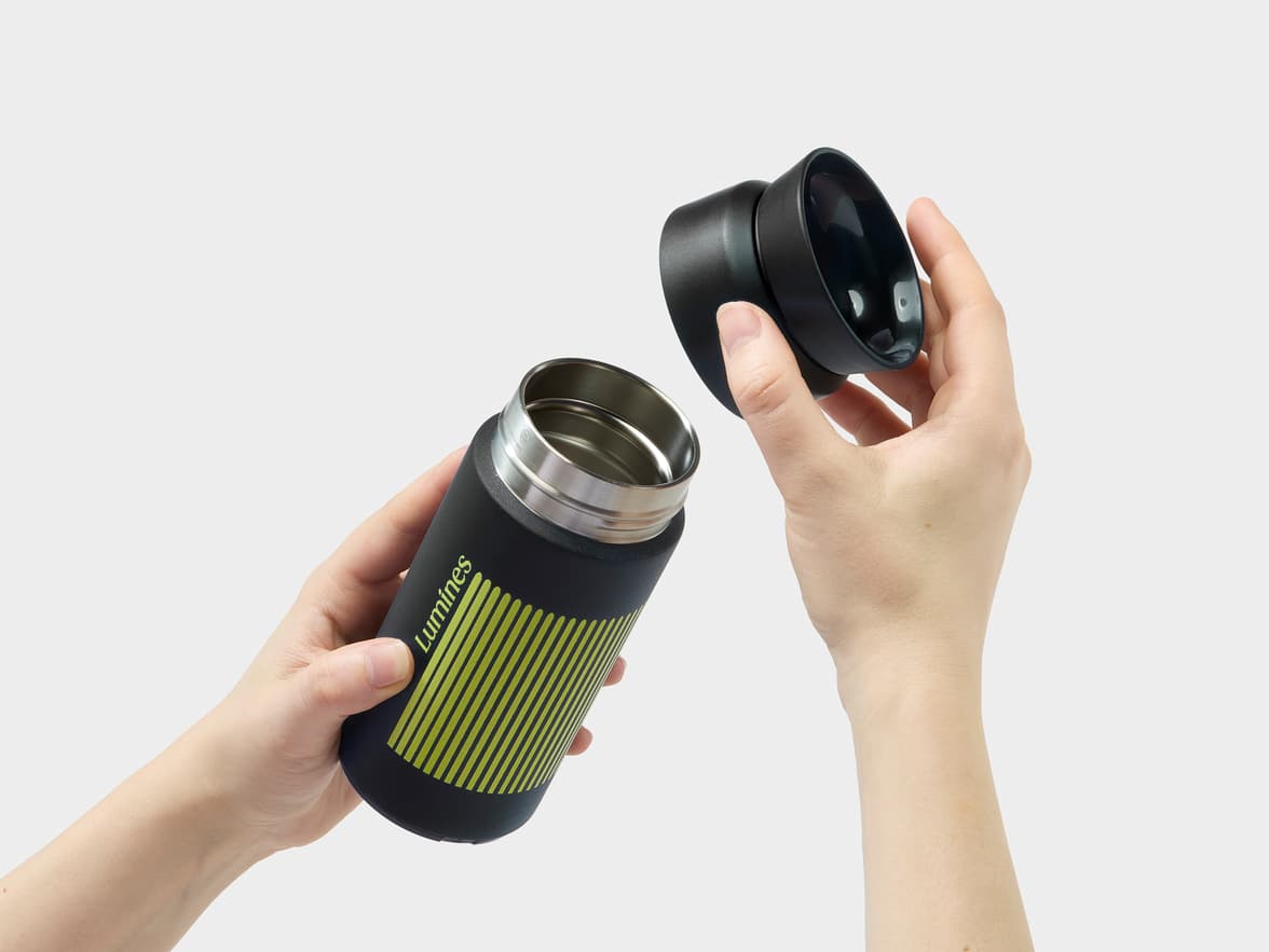 A person's hands open a black 'Lumines' brand travel mug with green stripes.
