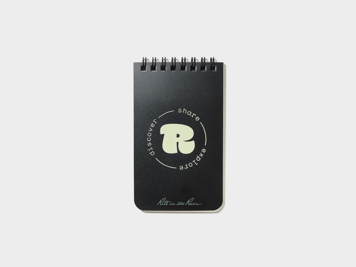 A black spiral-bound notebook featuring an "R" logo encircled by "discover share explore" and "Rite in the Rain" branding on its cover.