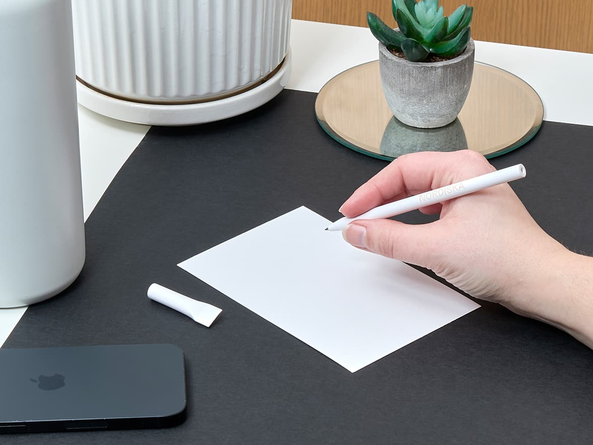 Hand holding White MOO Paper Pen on a desk with some paper, a phone and a Large Insulated Water Bottle