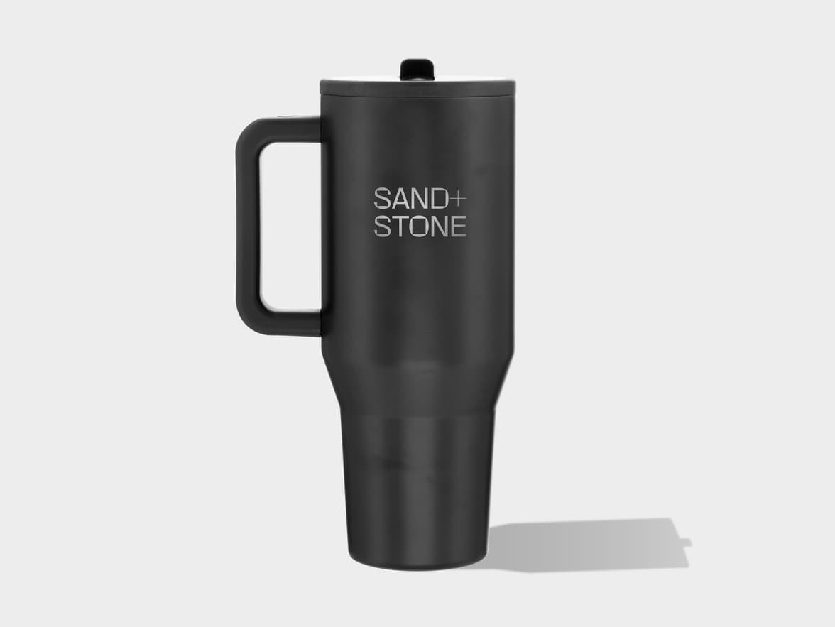 A black travel mug with a handle and lid, featuring the "SAND + STONE" logo on its side, stands on a light grey surface with a shadow cast to its right.
