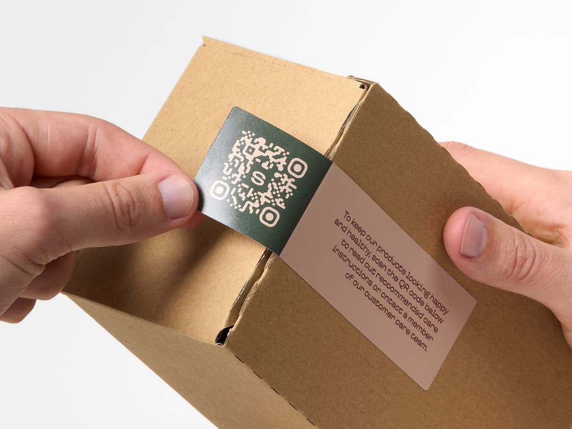 Hands placing a rectangular coated sticker with a QR code printed on it over the corner of a cardboard box.