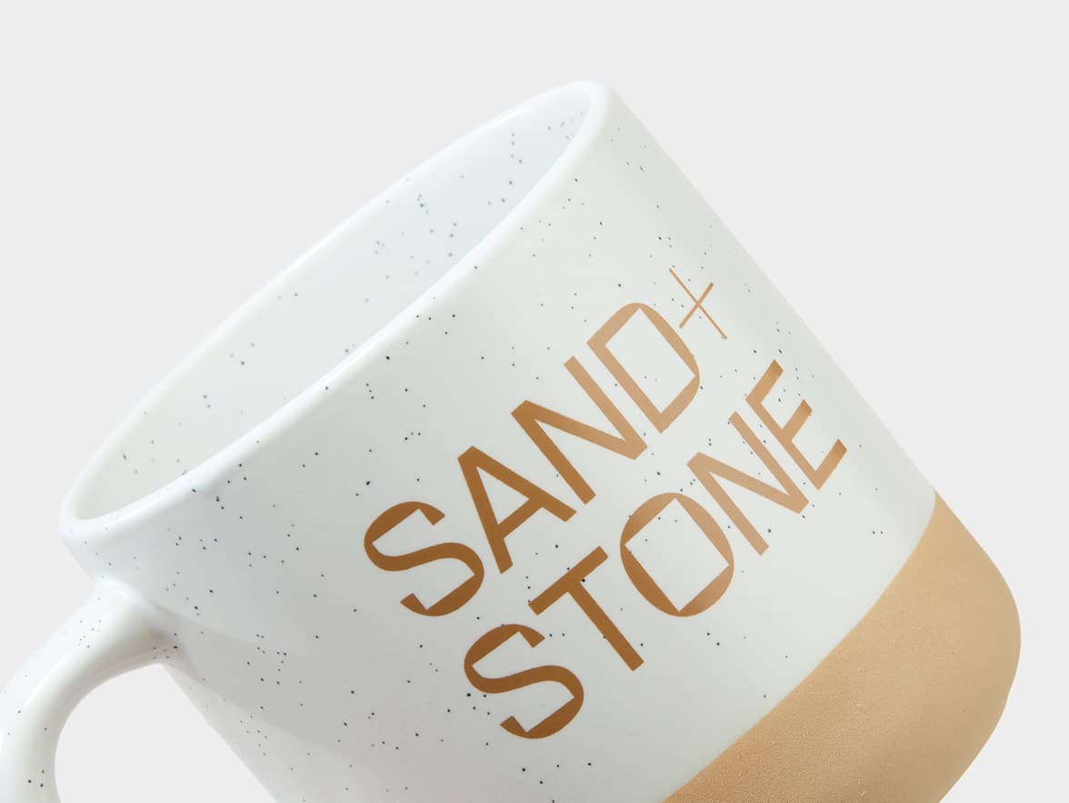 White speckled mug with a light brown base and "SAND + STONE" branding.
