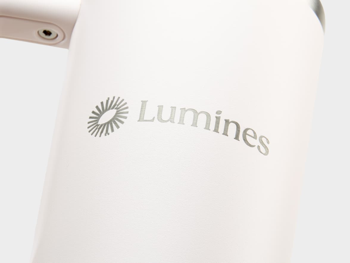 A close-up of a white textured tumbler with a metallic grey "Lumines" logo. The logo features a stylized sunburst icon to the left of the word "Lumines", which is subtly curved upwards.