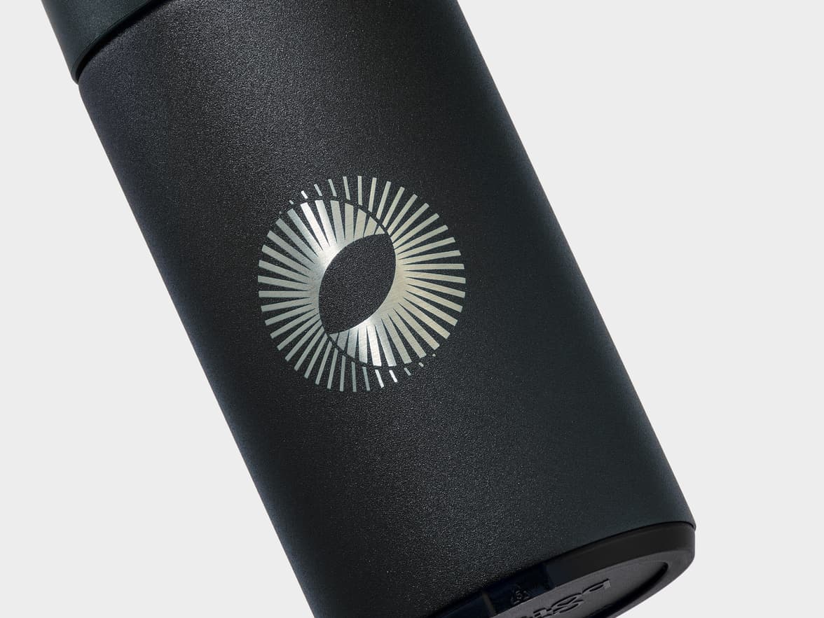 A close-up of a matte black, textured flask with a silver, circular, stylized logo featuring an oval surrounded by radiating lines.